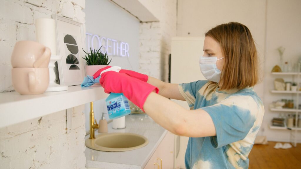 Person with pink gloves cleaning a shelf in a modern home, wearing a mask for safety during household chores.