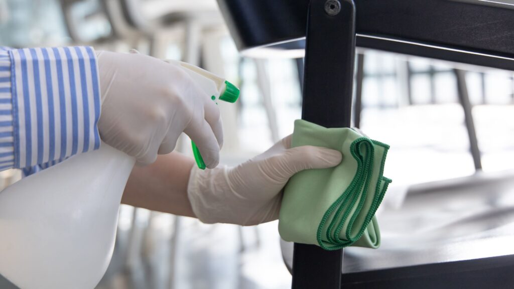 Person cleaning a chair using a spray bottle and green cloth, highlighting sanitization efforts in a clean environment.